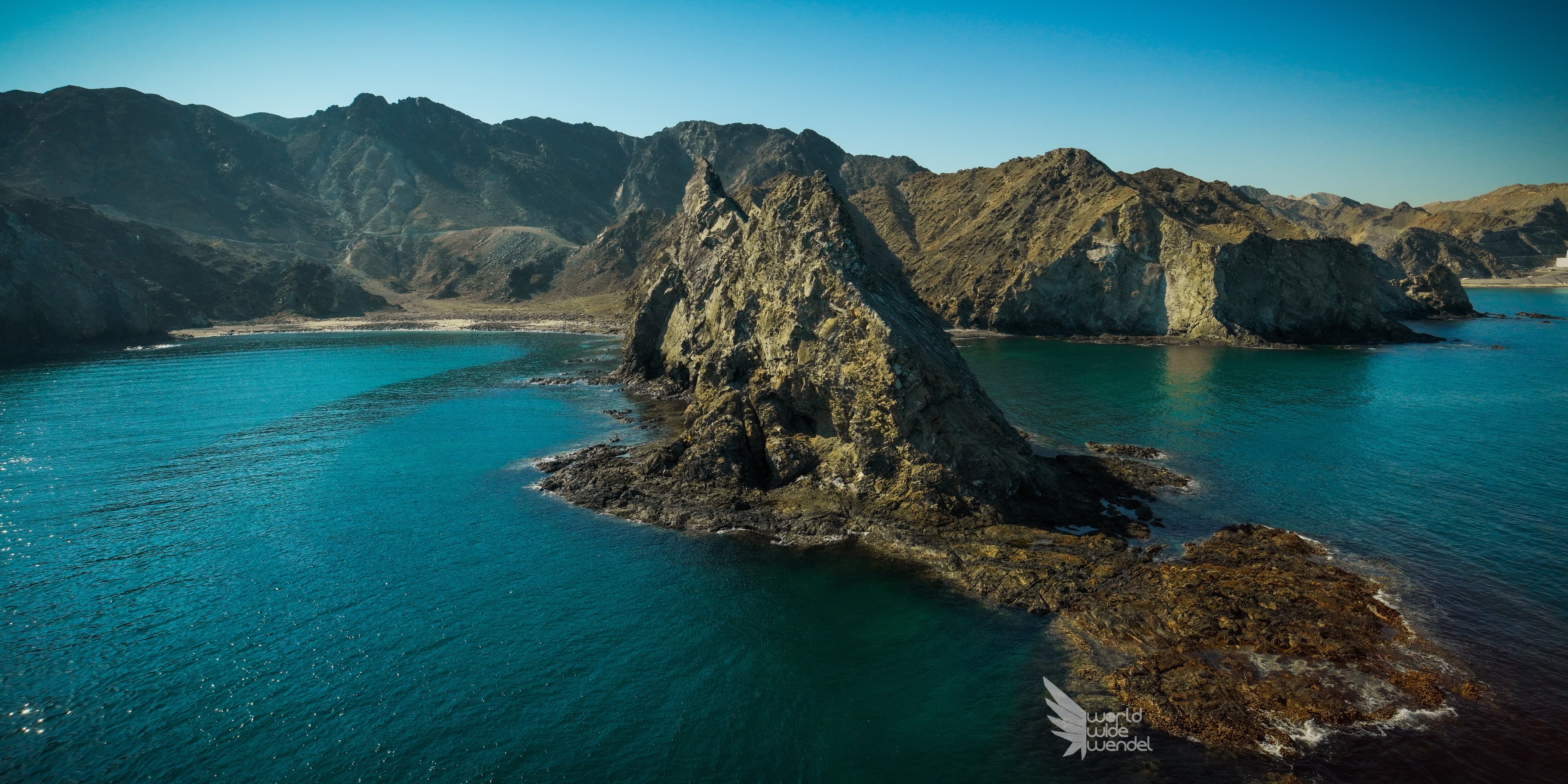 Luchtfoto van een rotsachtige kustlijn met een piramidevormige rots in turkoois water, Oman