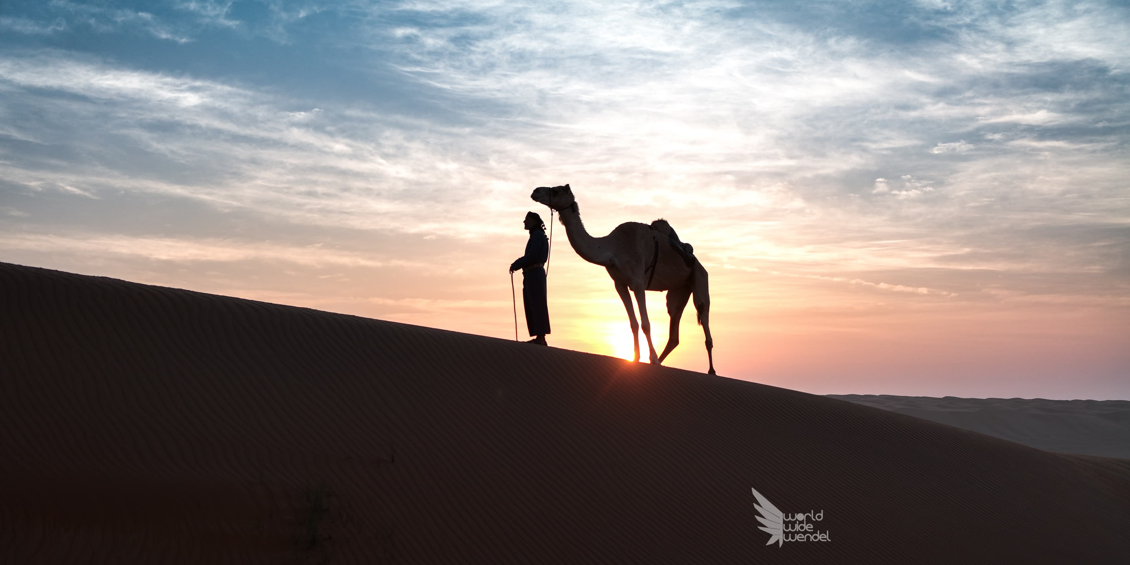 Silhouet van een man met een kameel op een duin bij zonsondergang